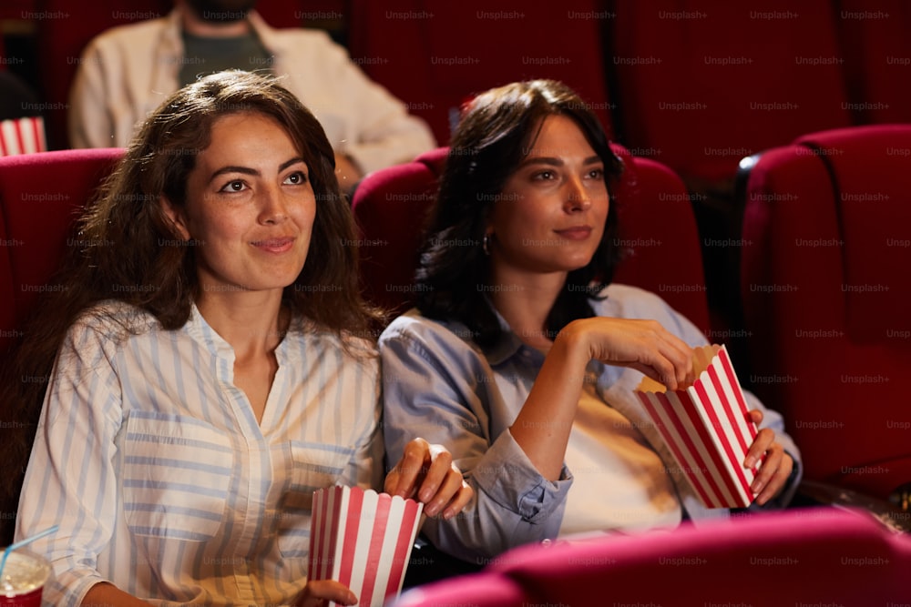 Portrait de deux jeunes femmes souriantes regardant un film au cinéma et mangeant du maïs soufflé assises sur des sièges en velours rouge, espace de copie