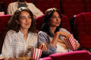 Portrait of two smiling young women watching movie in cinema and eating popcorn while sitting on red velvet seats, copy space