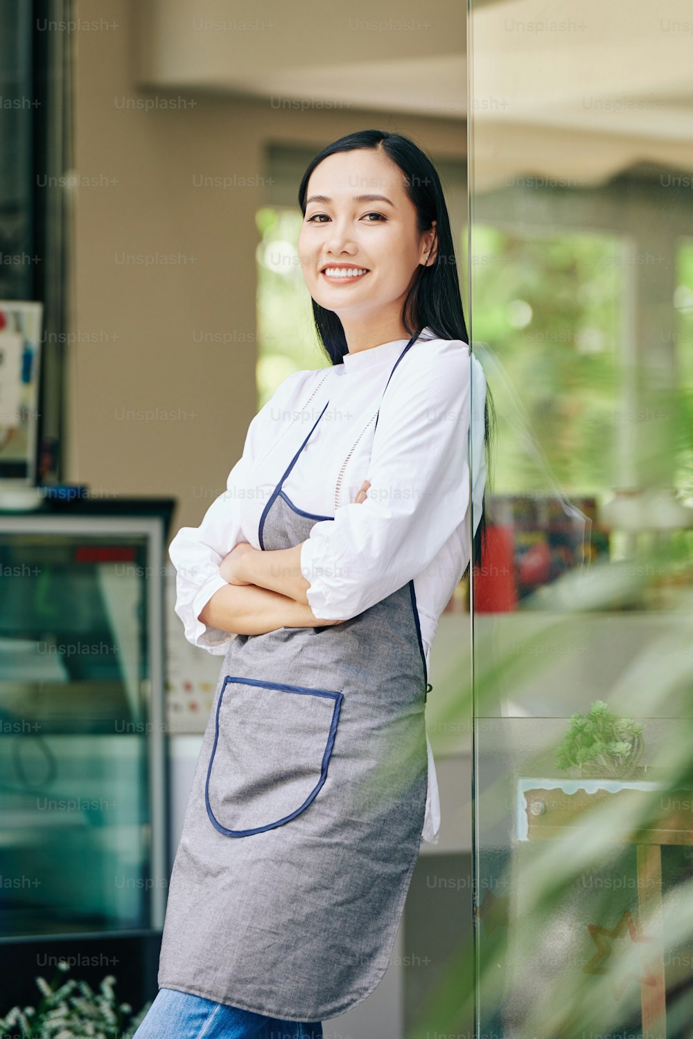 Beautiful young Asian waitress standing in doors of her coffeeshop and smiling at camera