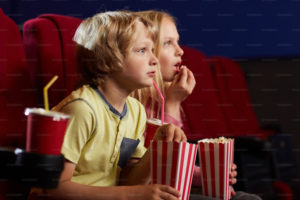 Side view portrait of two kids with open mouths watching movie in cinema theater and eating popcorn, copy space