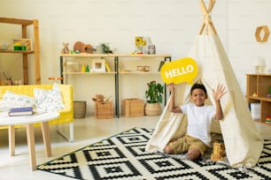 Cute African little boy waving hand and showing yellow speech bubble with greeting while sitting on the floor by white tent in living-room