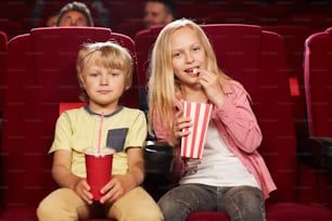 Front view portrait of two cute kids watching movie in cinema theater and eating popcorn, copy space