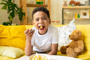 Joyful African little boy in t-shirt putting piece of food into his mouth while sitting on couch by table in living-room environment