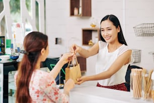 Feliz y joven dependienta vietnamita dando bolsa de papel con orden a la clienta