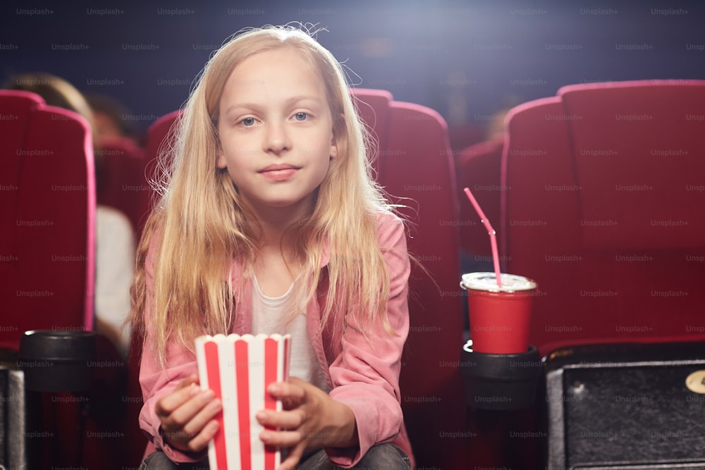 Front view portrait of blonde teenage girl looking at camera while holding popcorn cup in cinema theater, copy space
