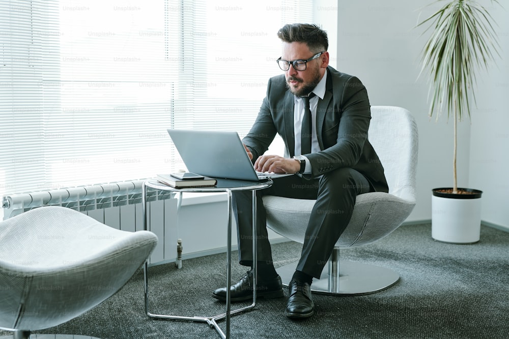 Hombre de negocios barbudo con traje elegante sentado en el sillón frente a la computadora portátil en la oficina y enviando correos electrónicos a un colega o socio comercial