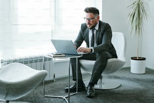 Bearded businessman in elegant suit sitting in armchair in front of laptop in office and sending e-mail to colleague or business partner