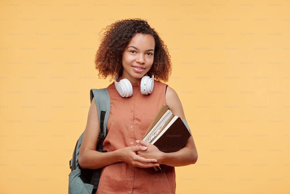 Portrait of smiling African-American student girl with headphones around neck holding books and satchel on back against bright background