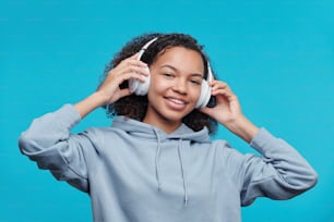 Porträt eines glücklichen hübschen schwarzen Mädchens im Kapuzenpullover, das drahtlose Kopfhörer anpasst, während es Musik vor blauem Hintergrund hört