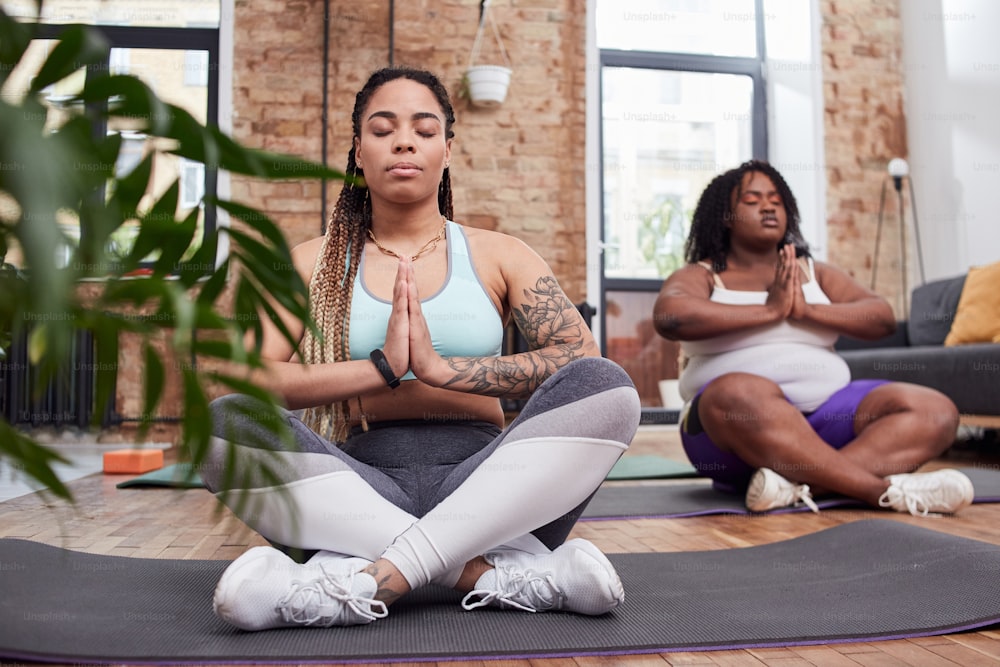 Senhoras relaxadas sentadas em posição de lótus e pressionando as palmas das mãos juntas enquanto praticam ioga no apartamento