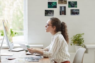 Ritratto di vista laterale della giovane donna moderna che tiene le fotografie mentre lavora al PC in ufficio bianco, spazio di copia