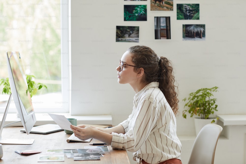 Side view portrait of modern young woman holding photographs while working at PC in white office, copy space