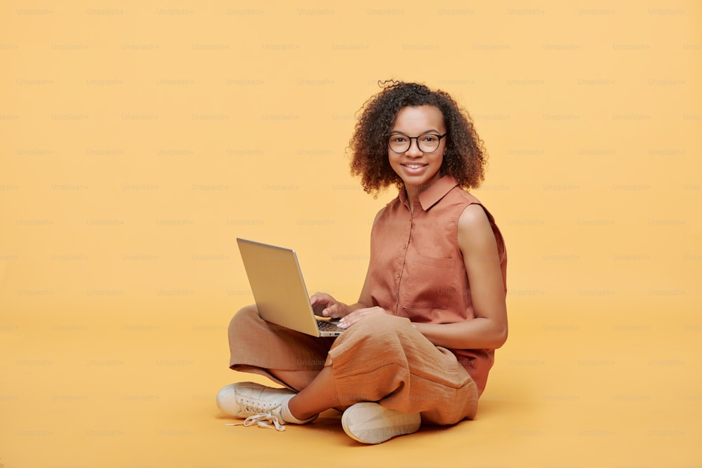 Portrait d’une étudiante afro-américaine souriante aux cheveux bouclés assise avec les jambes croisées et utilisant un ordinateur portable sur fond jaune