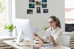 Retrato de una joven moderna sosteniendo fotografías revisando para publicar mientras trabaja en PC en oficina blanca, espacio de copia