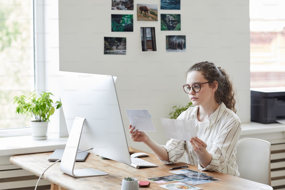 Portrait of modern young woman holding photographs reviewing for publishing while working at PC in white office, copy space