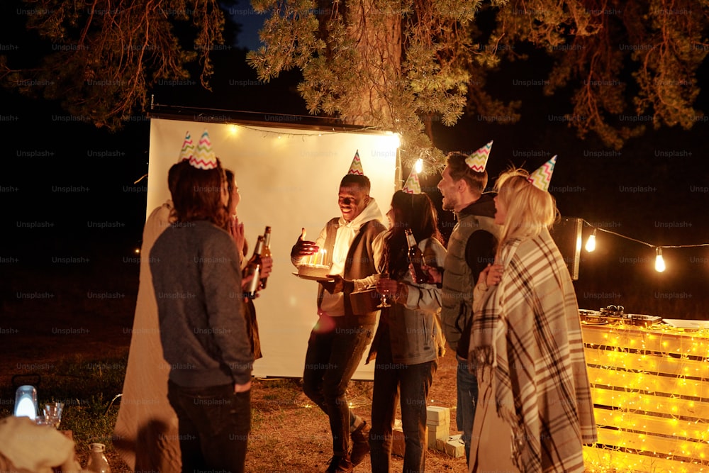 Happy young African man keeping hand by candles on birthday cake while standing among friends in caps toasting with bottles of beer