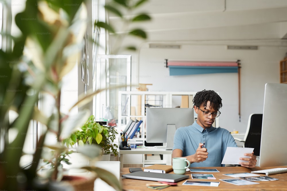 Portrait of creative African-American man reviewing photographs while working on editing and publishing in modern office, copy space