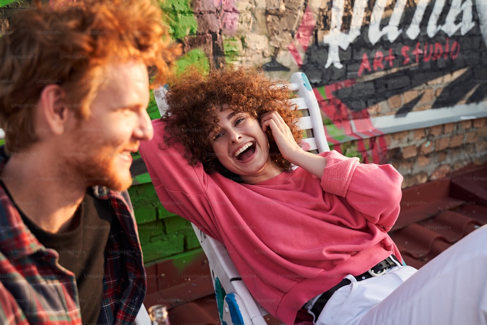 Happy curly female is lying on chaise lounge and laughing while communicating with boyfriend in open air