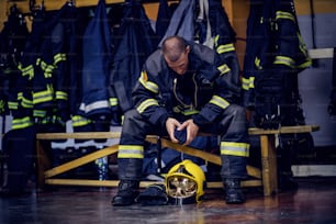Joven y atractivo bombero con uniforme protector sentado en la estación de bomberos y esperando a otros bomberos. Está preparado para la acción.