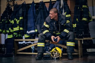 Junger attraktiver Feuerwehrmann in Schutzuniform, der in der Feuerwache sitzt und auf andere Feuerwehrleute wartet. Er ist bereit zu handeln.