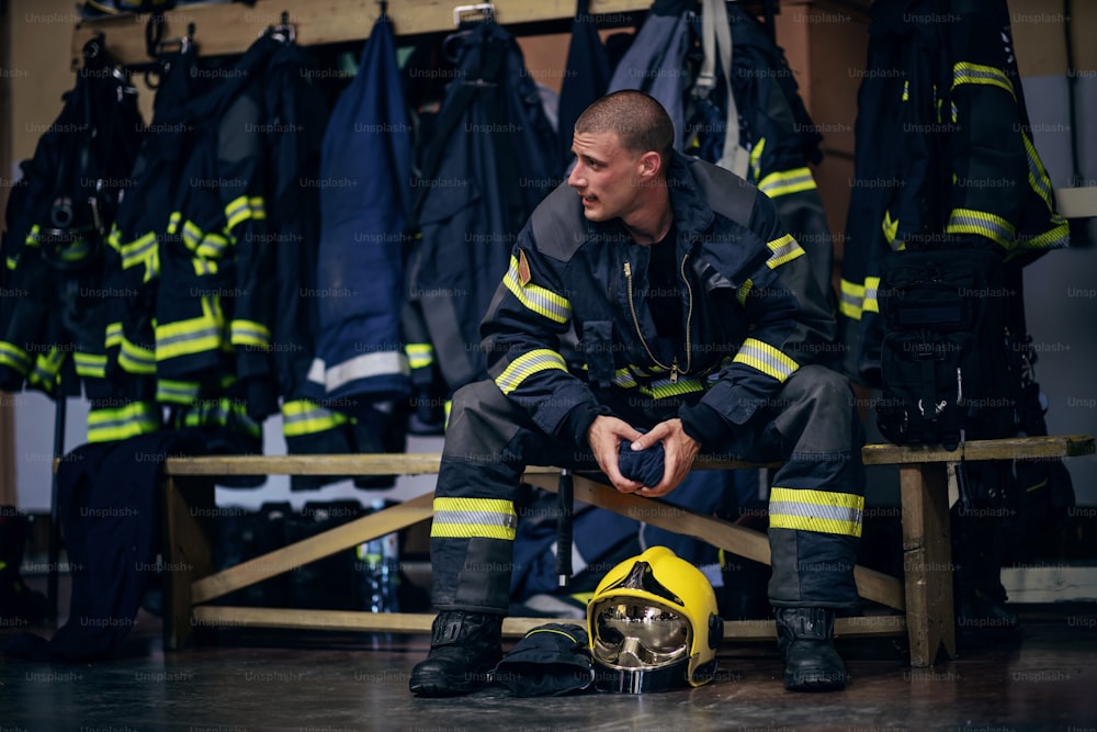 Joven y atractivo bombero con uniforme protector sentado en la estación de bomberos y esperando a otros bomberos. Está preparado para la acción.