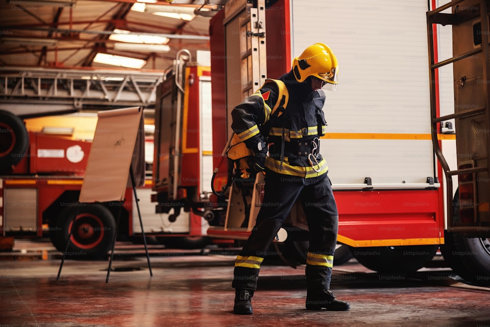 Figura intera di coraggioso vigile del fuoco in piedi nella stazione dei pompieri in uniforme protettiva e pronto per l'azione.
