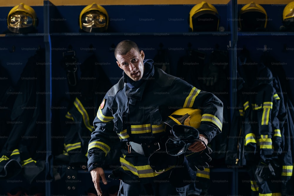 Valiente joven y atractivo bombero con uniforme protector que sostiene el casco debajo de la axila después de la acción mientras está de pie en la estación de bomberos.