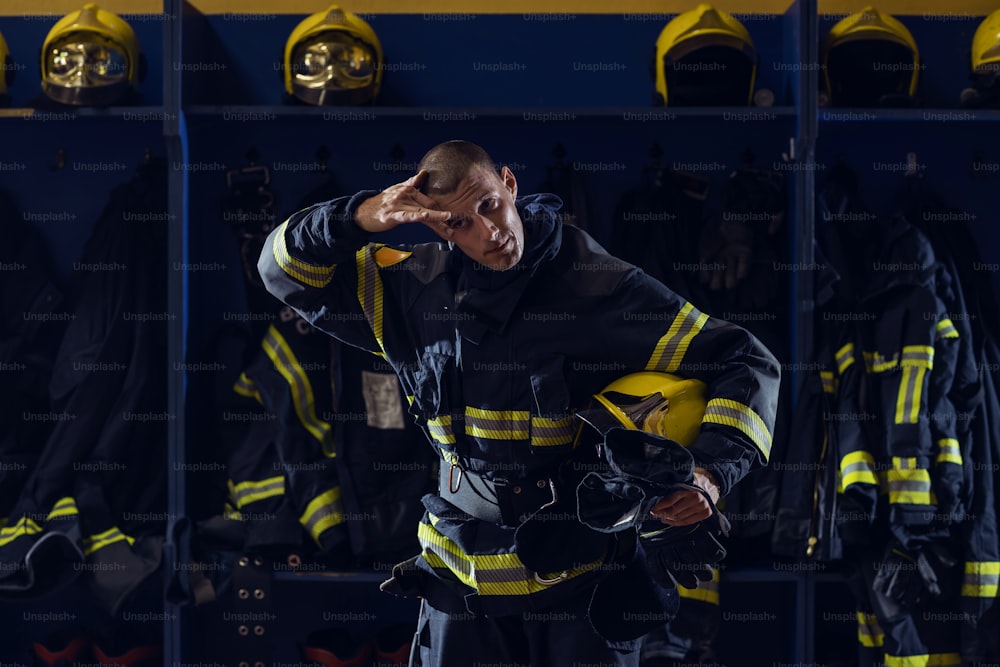 Valiente joven y atractivo bombero con uniforme protector, con casco debajo de la axila limpiando el sudor de la frente y descansando después de la acción mientras est�á de pie en la estación de bomberos.