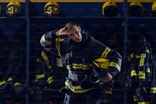 Brave young attractive fireman in protective uniform, with helmet under armpit wiping sweat from forehead and resting after action while standing in fire station.