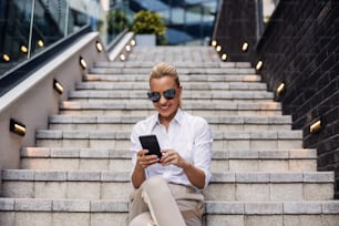 Sonriente hermosa rubia mujer de negocios de moda sentada en las escaleras frente al centro de negocios y usando un teléfono inteligente.