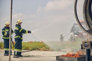 Vue latérale de deux pompiers en uniforme de protection et casques jaunes éclaboussant l’eau du tuyau