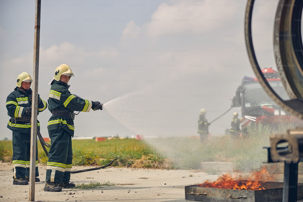 Vue latérale de deux pompiers en uniforme de protection et casques jaunes éclaboussant l’eau du tuyau