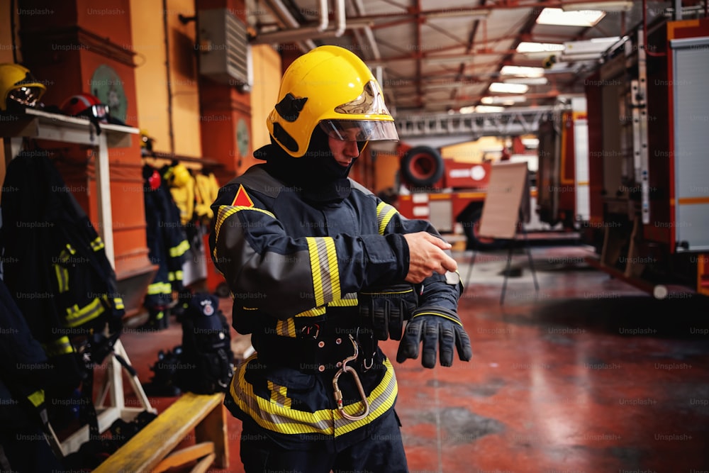 Fireman putting on protective uniform and preparing for action while standing in fire station.