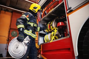 Firefighter in protective uniform with helmet on head checking on hoses before intervention while standing in fire station.