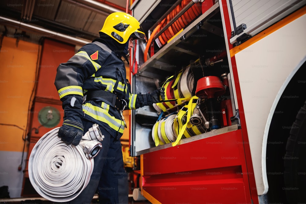 Vigile del fuoco in uniforme protettiva con casco sulla testa che controlla i tubi prima dell'intervento mentre si trova nella stazione dei pompieri.