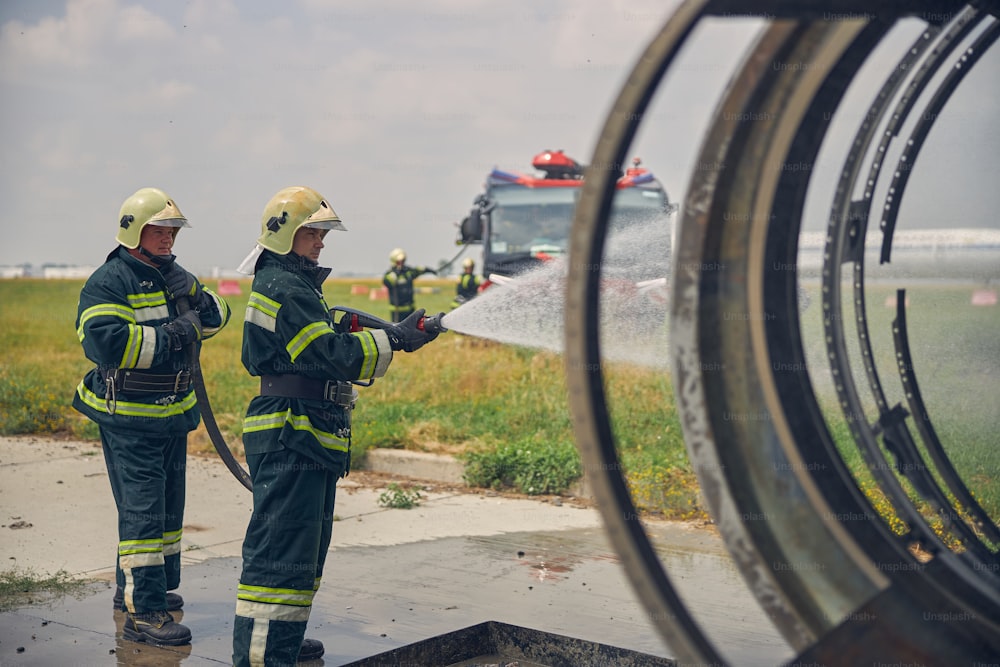 Deux pompiers tenant un tuyau d’arrosage à la main debout devant une construction métallique