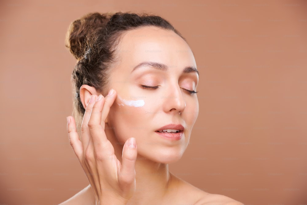 Young gorgeous woman applying facial cream or serum on undereye area while taking care of her skin in the morning or before sleep