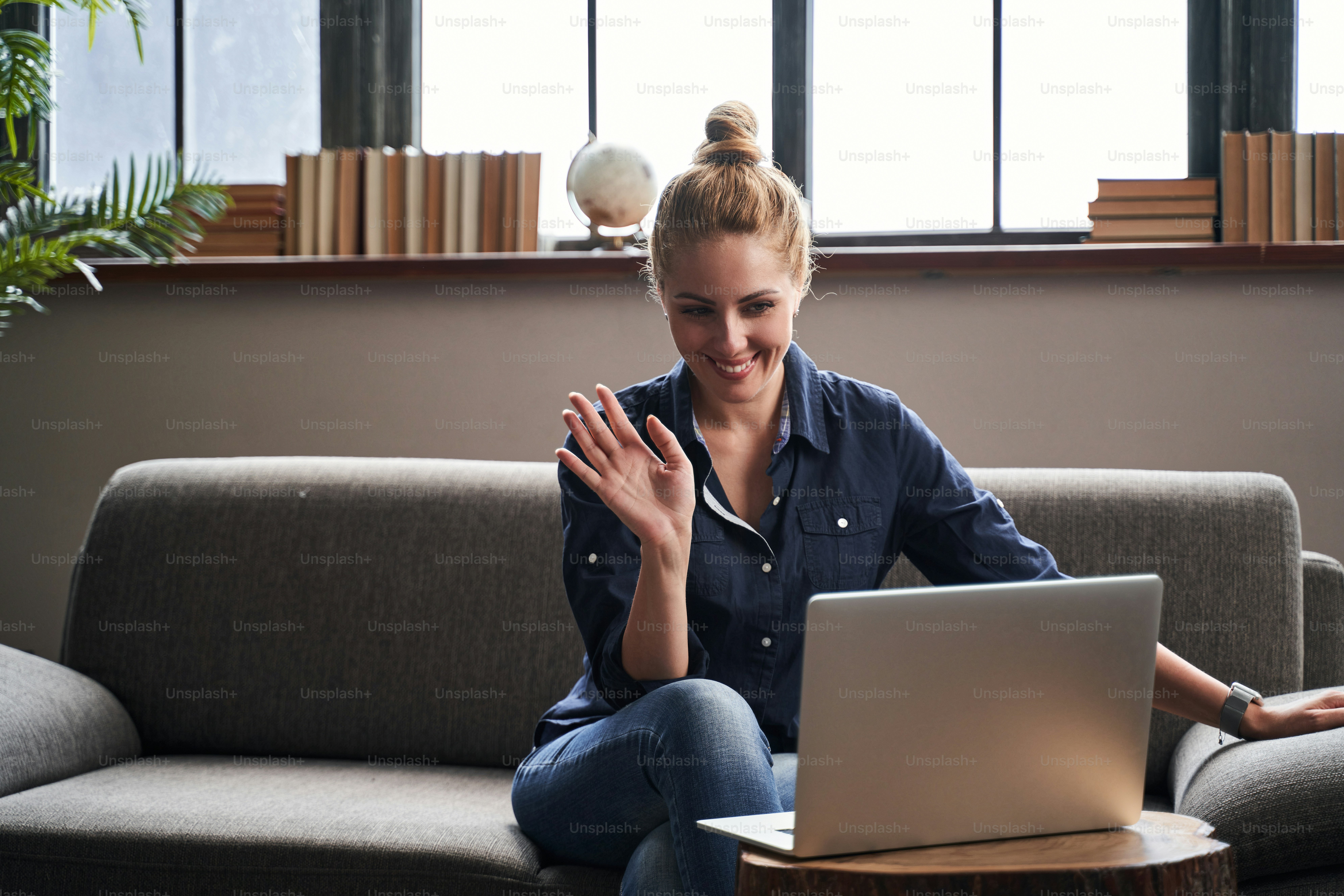 Cheerful young lady sitting on a sofa with a laptop and putting her hand up in front of a web camera