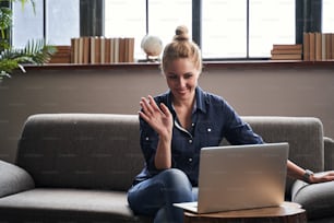 Cheerful young lady sitting on a sofa with a laptop and putting her hand up in front of a web camera