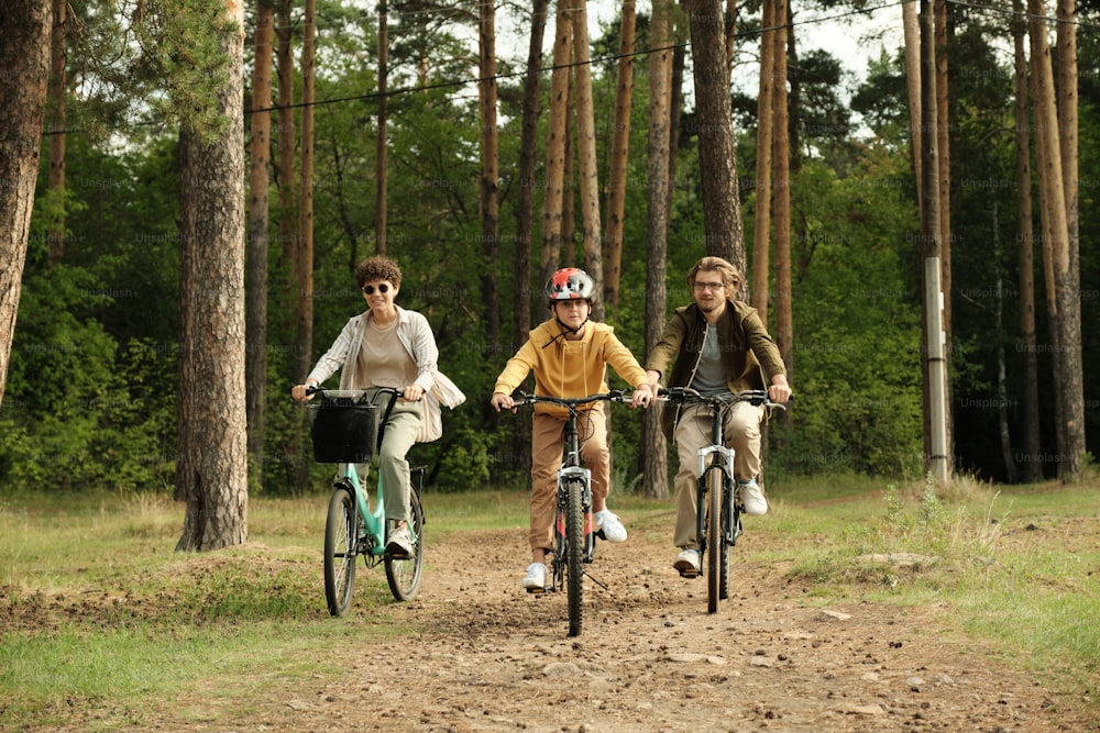 Happy active family of young husband, wife and son riding bicycles along forest path against pinetree trunks and other trees on summer day