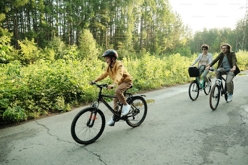 Giovane coppia e il loro figlio in bicicletta lungo la strada in ambiente naturale contro alberi verdi e cespugli durante il fine settimana estivo