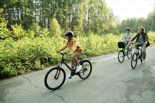Young couple and their son riding bicycles along road in natural environment against green trees and bushes on summer weekend