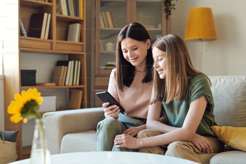 Young smiling female scrolling in smartphone through photos while showing them to her cute blond teenage daughter in living-room