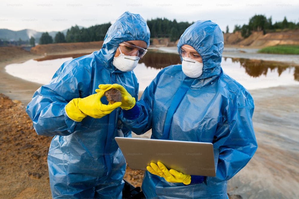 Young scientist in protective coveralls giving stone to colleague with laptop while both working over scientific investigation of polluted area