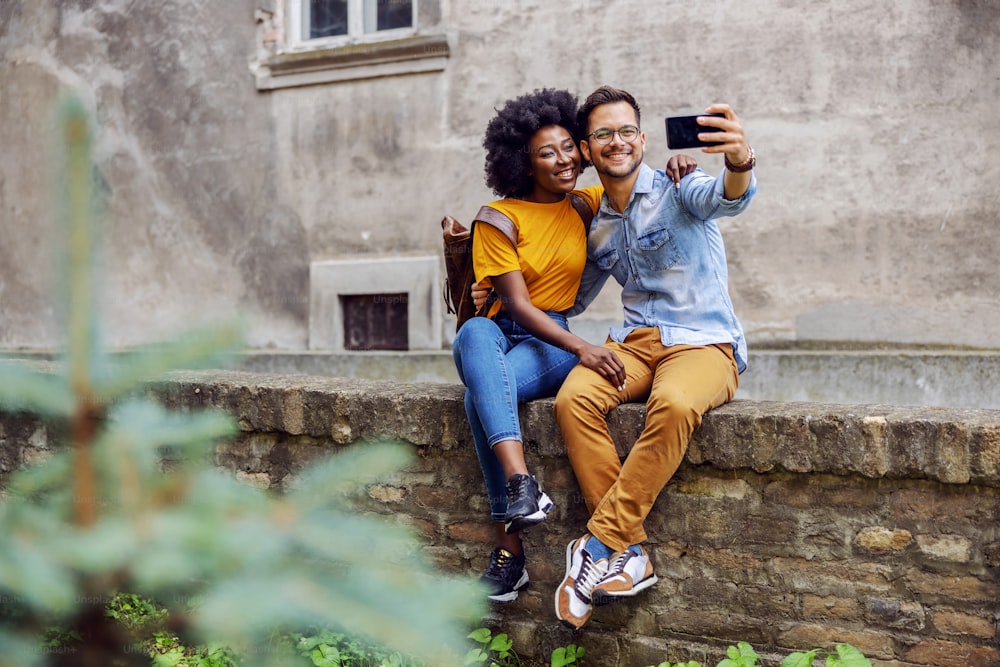Young multiracial couple sitting on a wall and taking selfie.