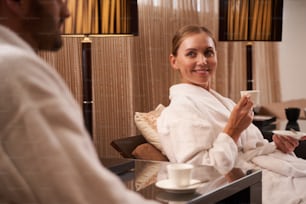 Gladsome woman in a white bathrobe kindly smiling to a man while holding a cup of coffee