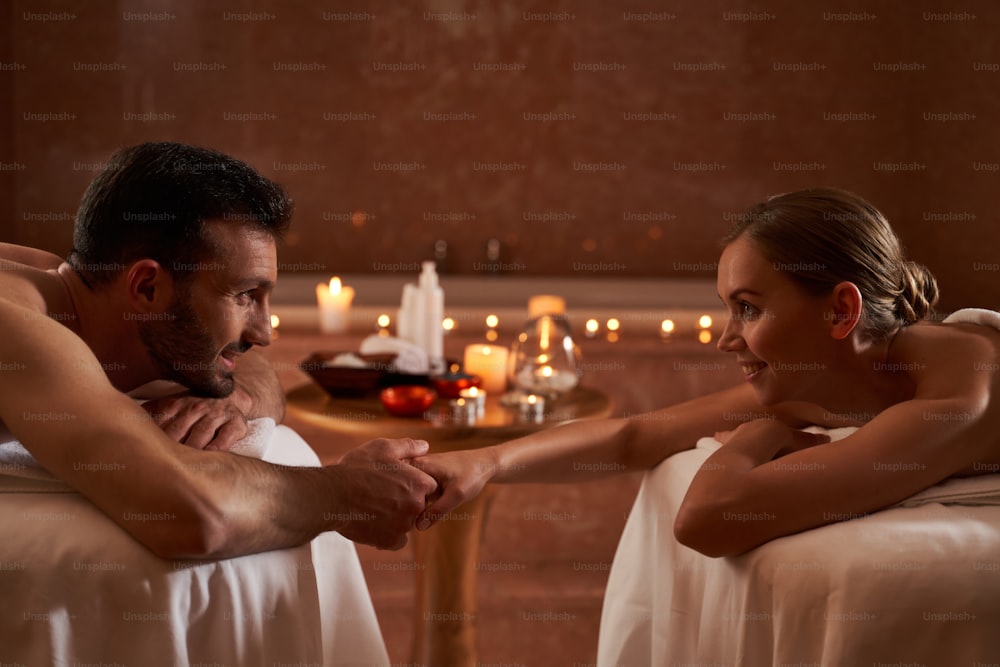 Happy young couple lying on massage tables and smiling to each other while holding hands. Light of burning candles on the background