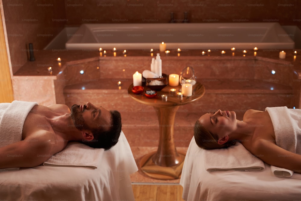 Hermosa pareja relajándose con los ojos cerrados y disfrutando del ambiente de la sala de masajes con velas encendidas