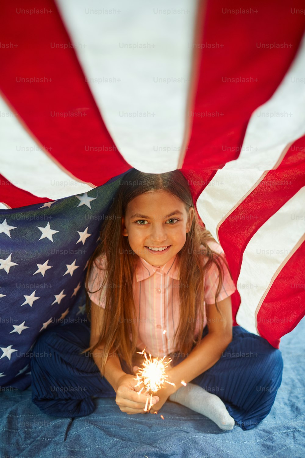 Retrato de alto ángulo de una linda chica sosteniendo luces brillantes sonriendo a la cámara cubierta por la bandera estadounidense, espacio de copia