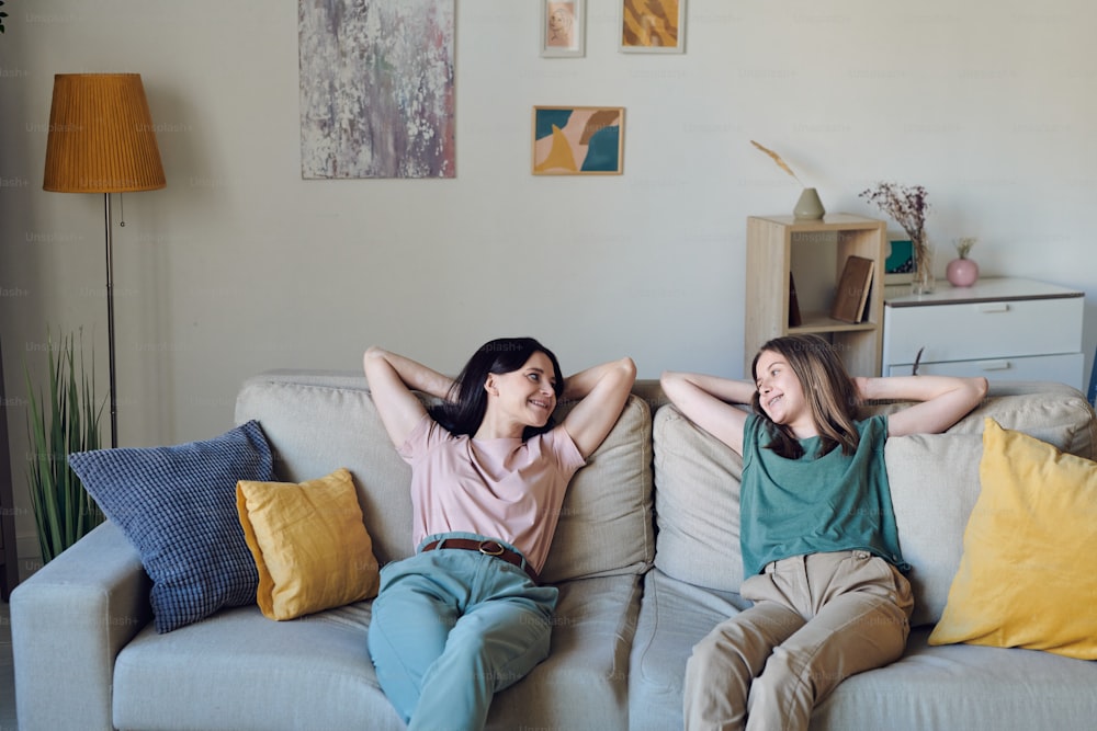 Cheerful and relaxed mother and daughter in casualwear looking at each other while sitting on soft couch and chatting in home environment